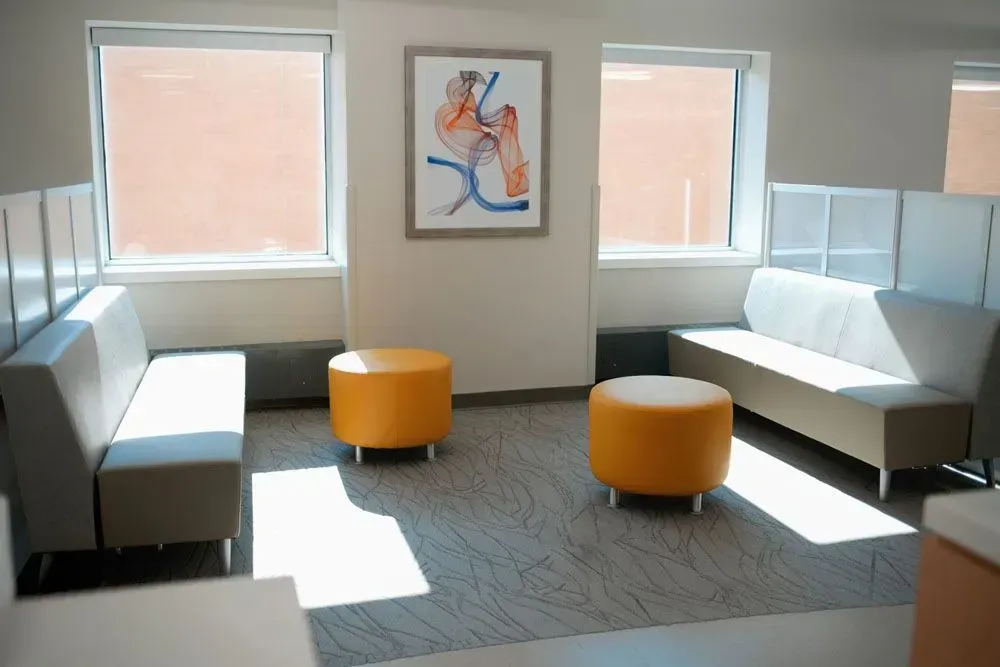 Waiting room with two beige sofas, two orange ottomans, and a framed anatomical art piece between two windows.