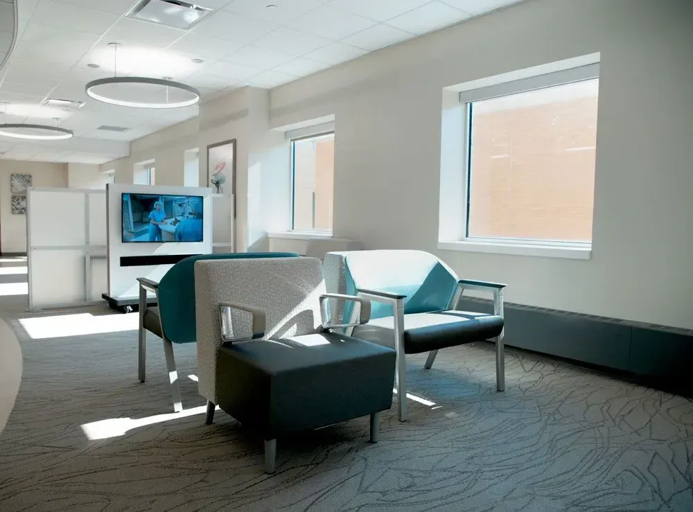 A modern waiting area with light-colored chairs, carpet, and a large window. A television screen is visible.