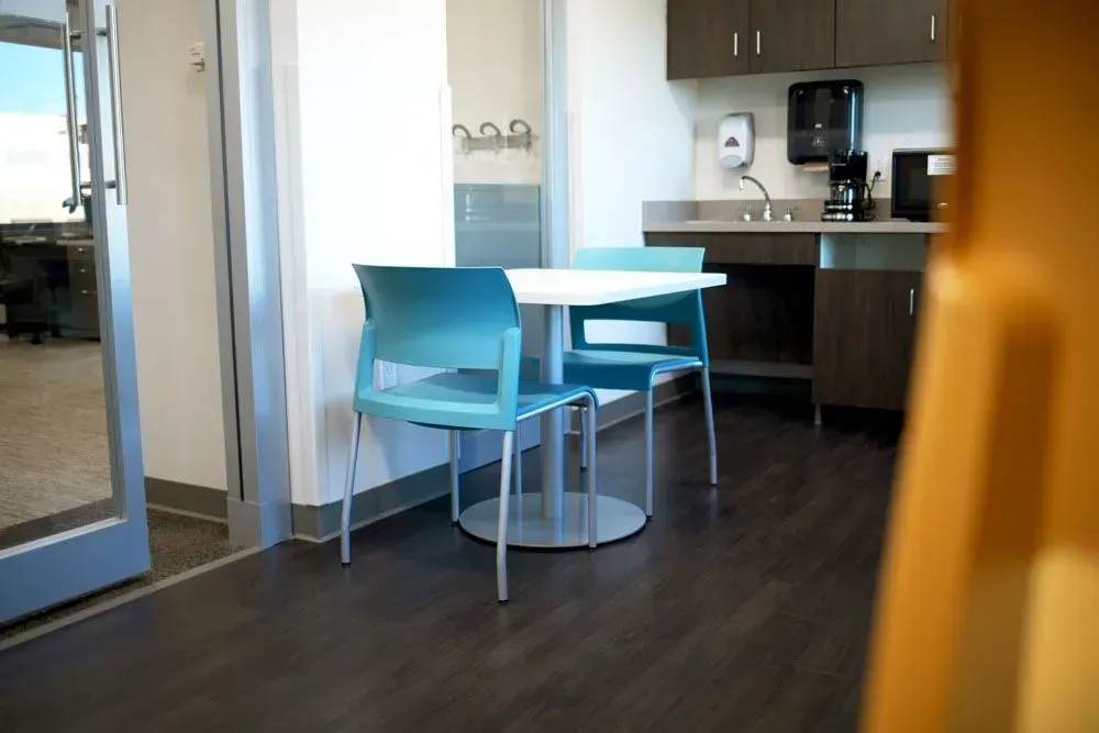 Small break room with table, chairs, sink, cabinets, coffee maker; light blue and dark brown colors.