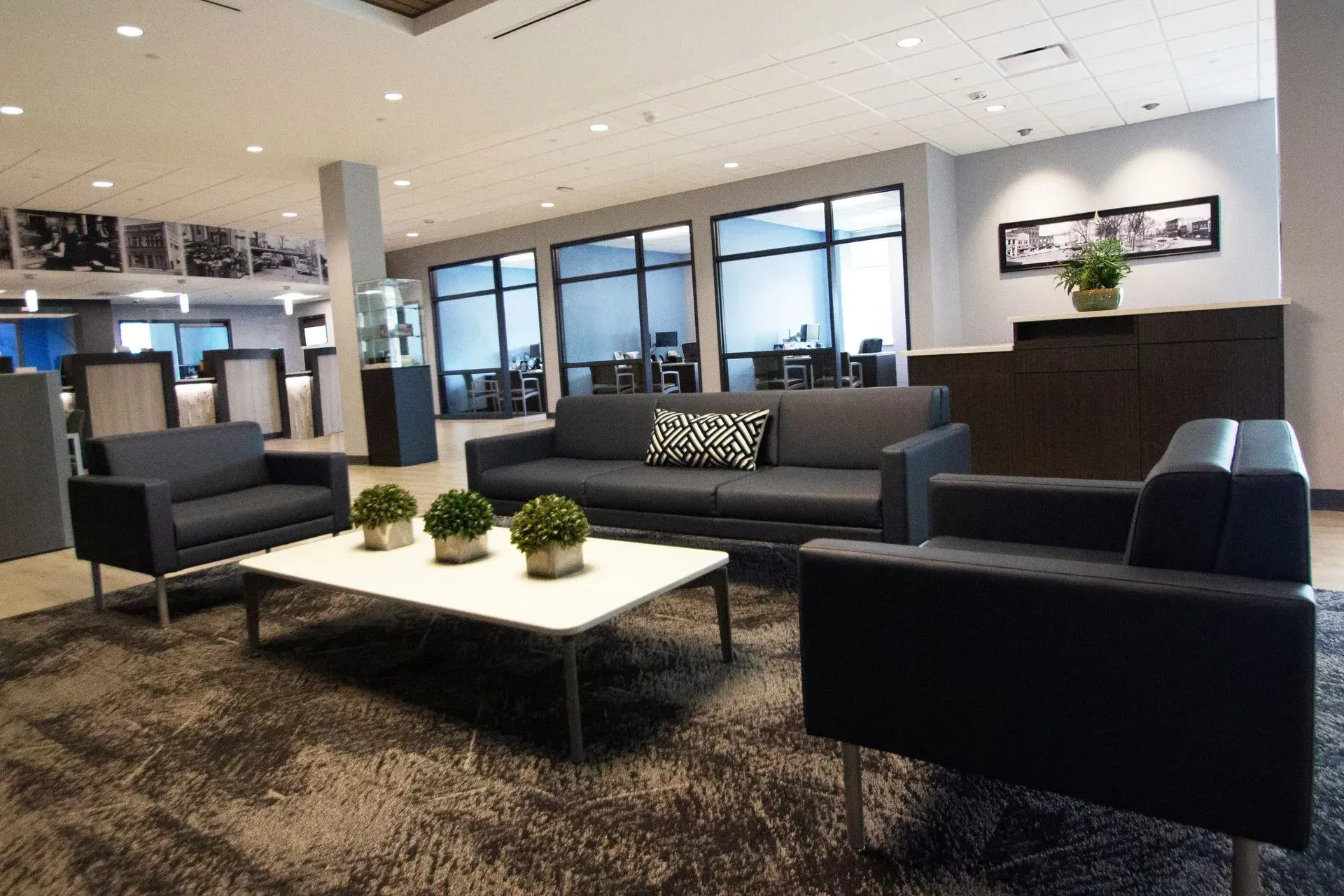 Lounge area with gray sofa, armchairs, white coffee table, and large windows looking into an office.