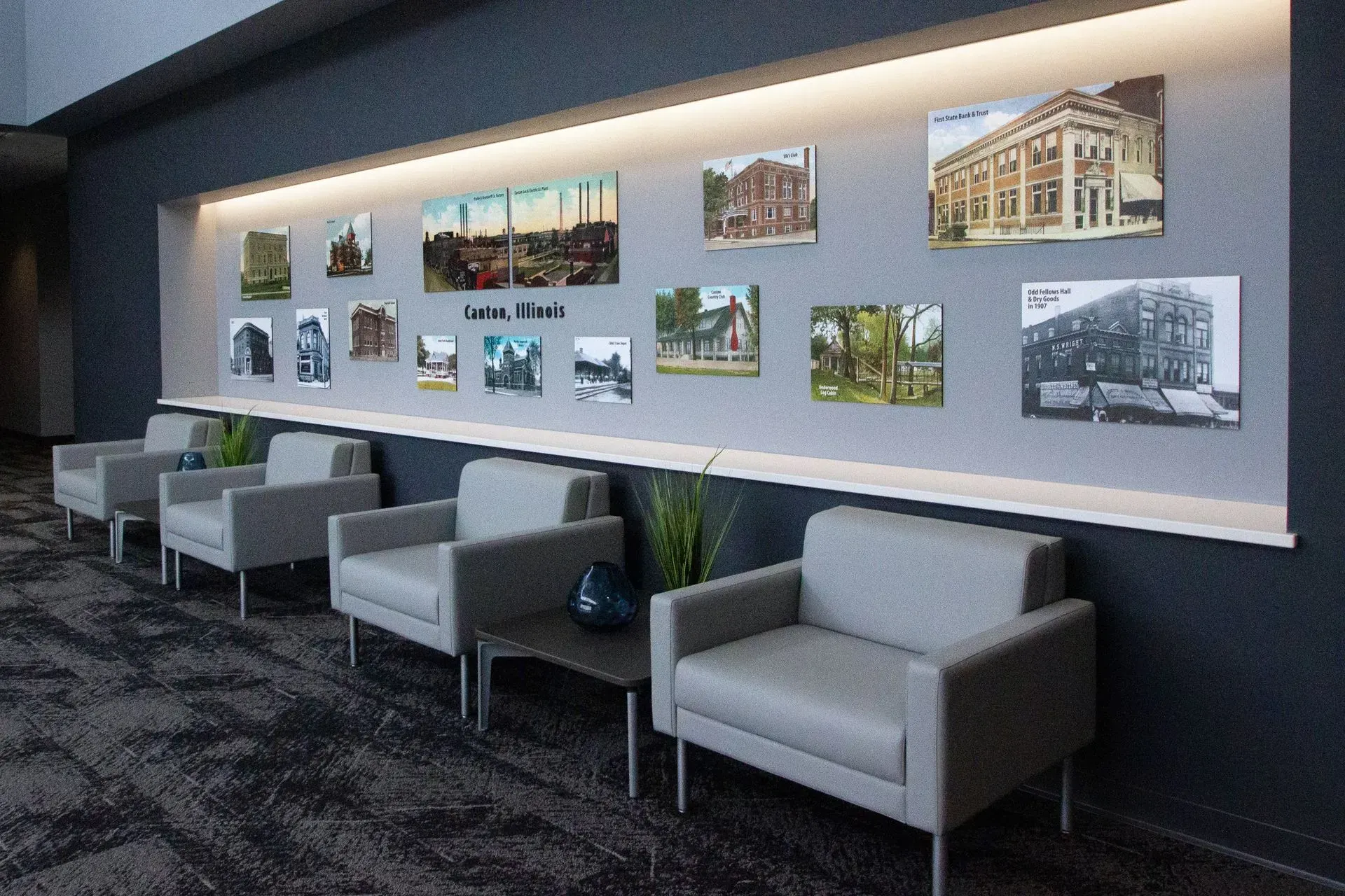 Lounge area with gray armchairs and a display of historical photos, illuminated against a dark wall.
