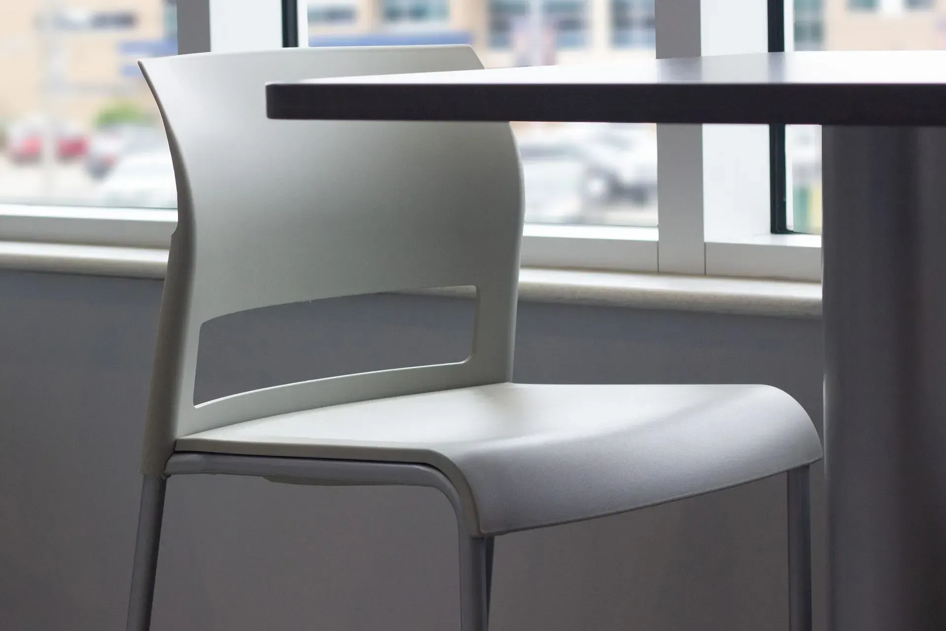 White chair with cut-out detail under a dark tabletop, beside a window with a blurred outdoor view.