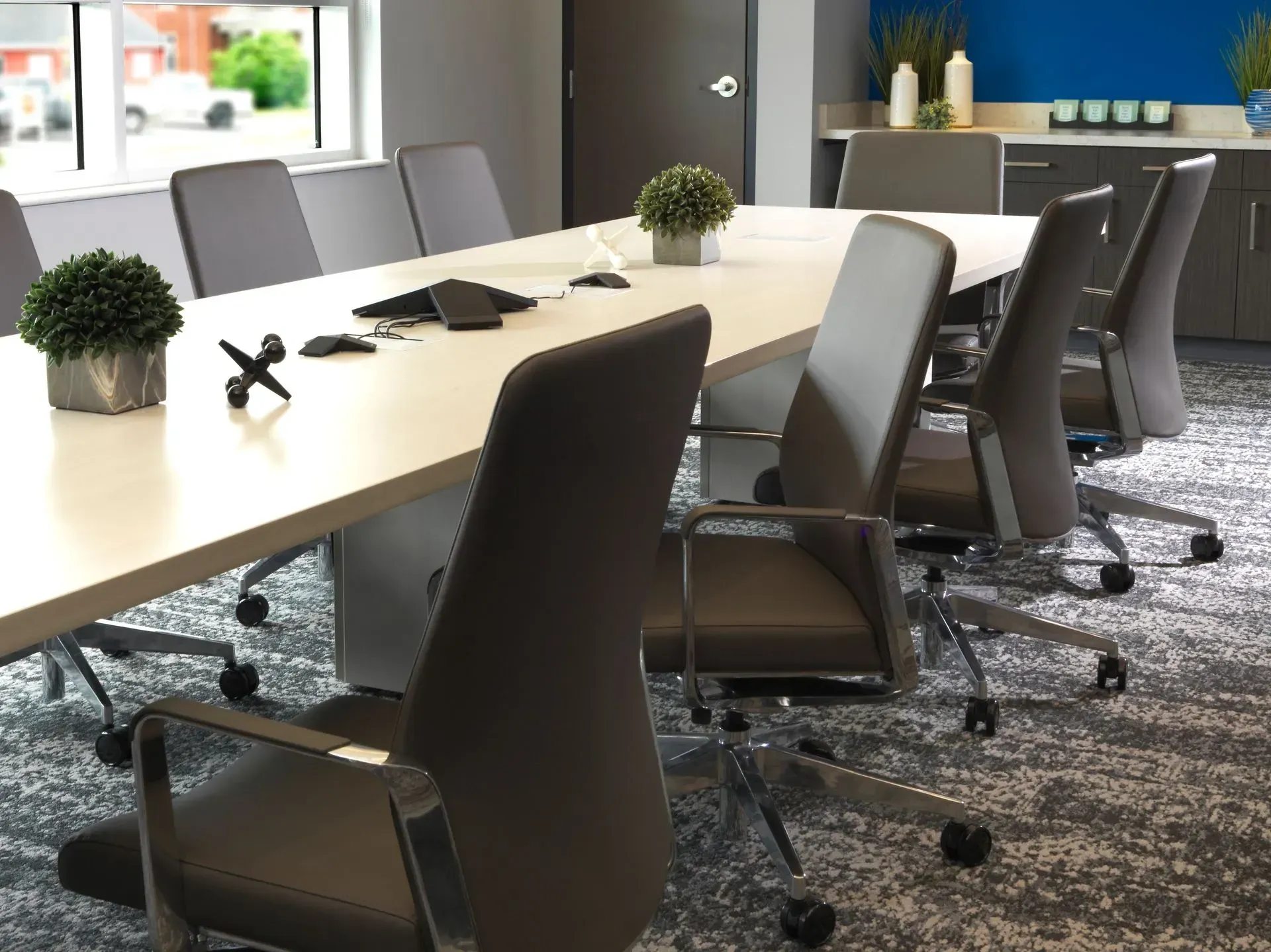 Conference room with long white table, gray chairs, small plants, and a window.