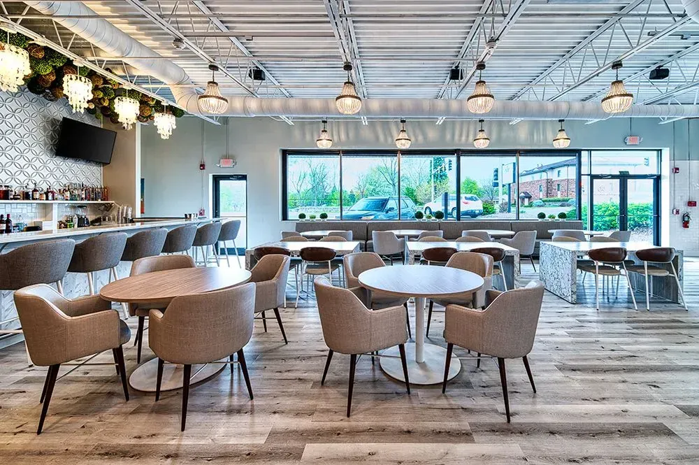 Restaurant interior with tables and chairs, bar, large windows, and decorative lighting.