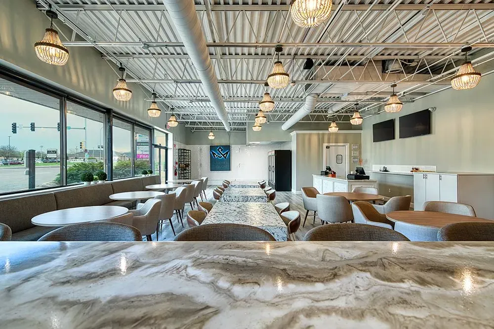 Interior view of a modern cafe with tables, chairs, and decorative lighting. Large windows offer an outside view.