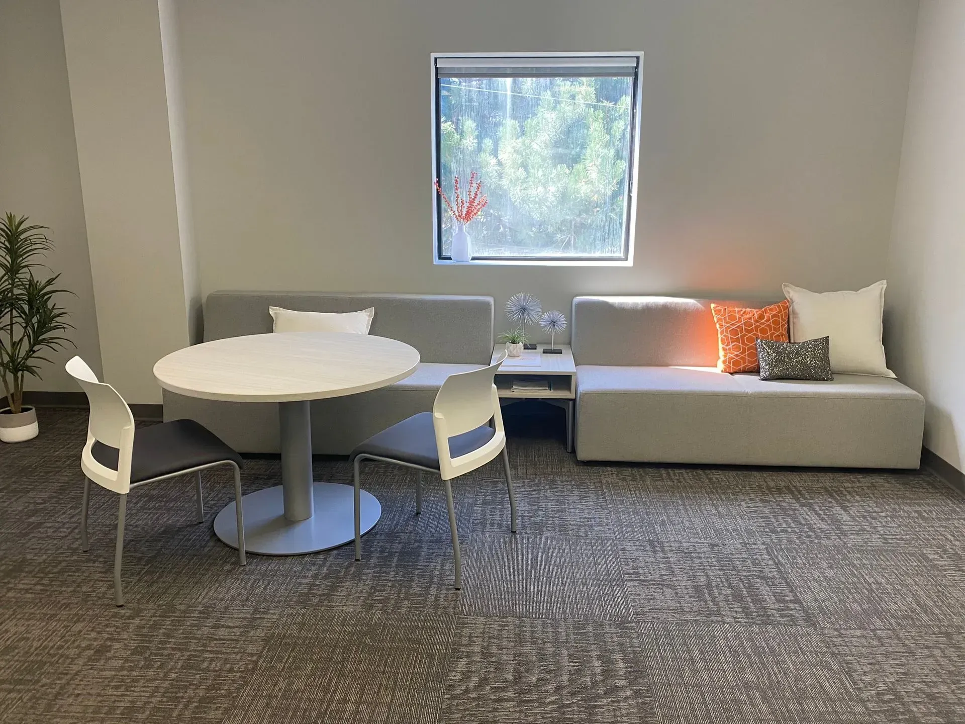 Cozy office lounge with gray seating, round table, small window, and pillows.