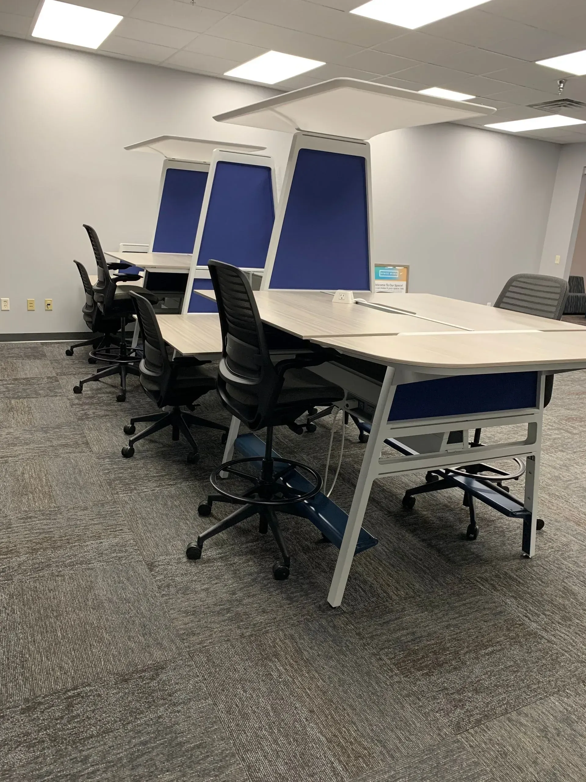 Office space with long desks, blue panels, and black chairs. Gray carpet, white walls, and bright ceiling lights.