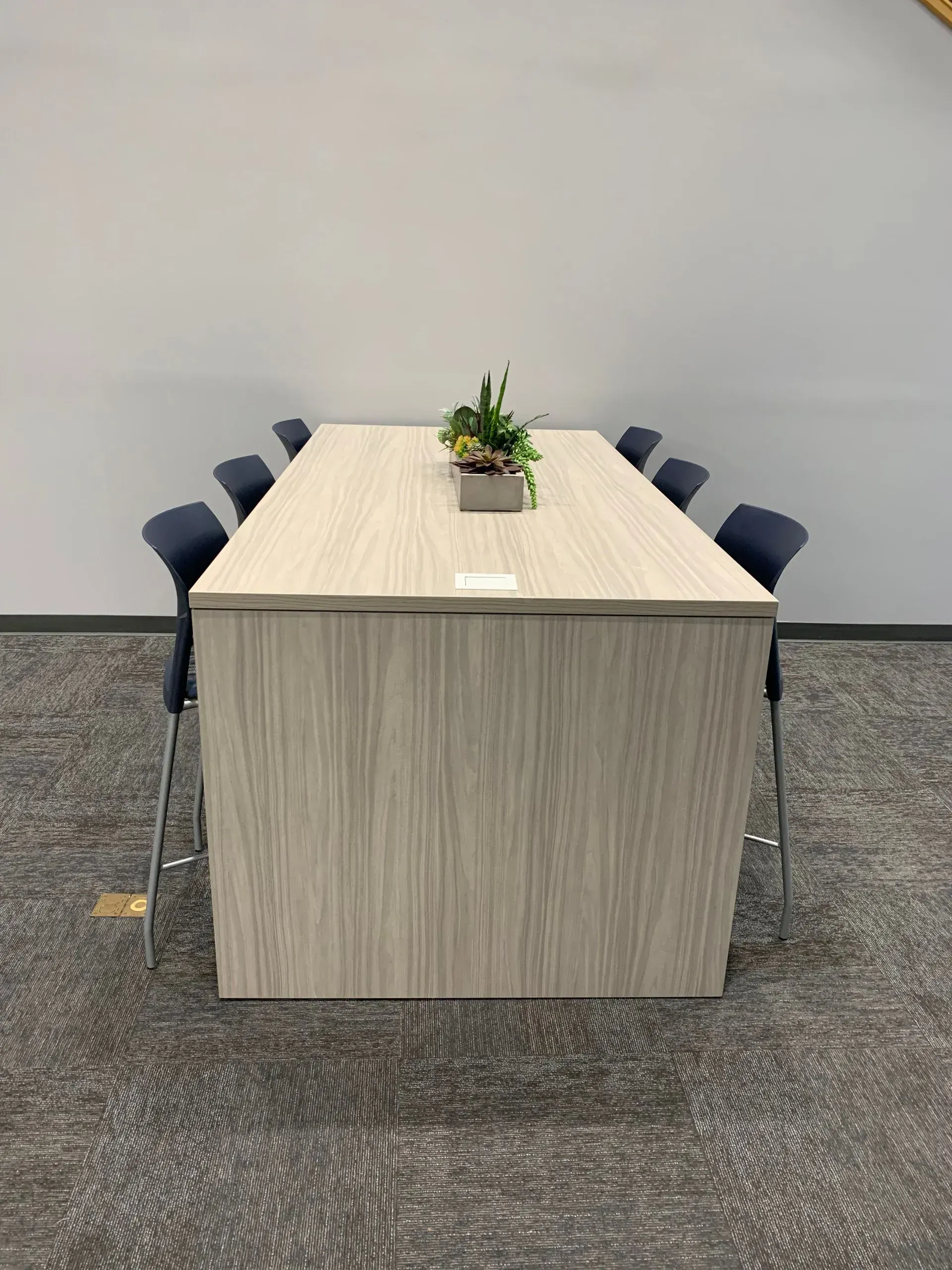 A rectangular, light wood-grain table with eight dark blue barstools. A plant sits in the center. Carpeted floor.