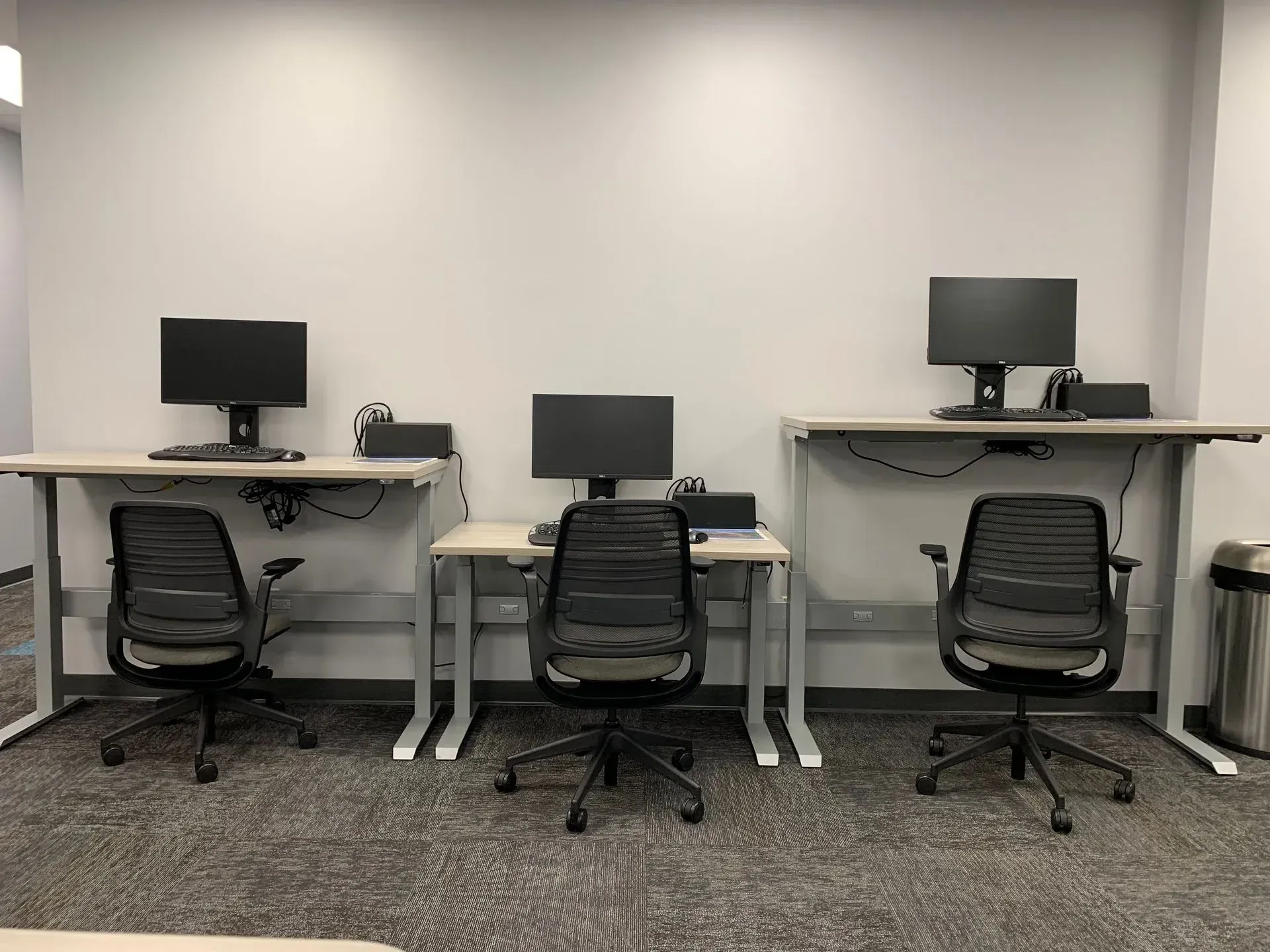 Three computer workstations with black monitors, chairs, and adjustable desks against a white wall.