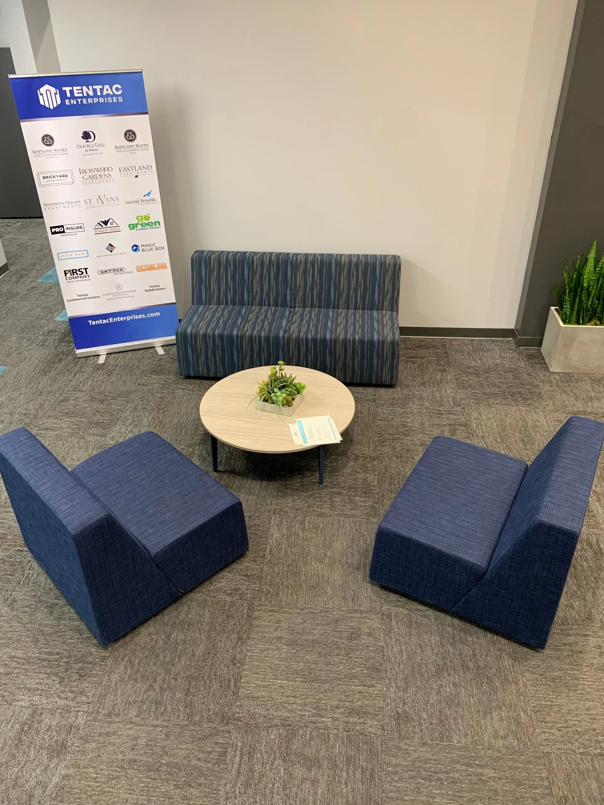 Lounge area with blue patterned furniture, a round table, and a company banner in a modern office.