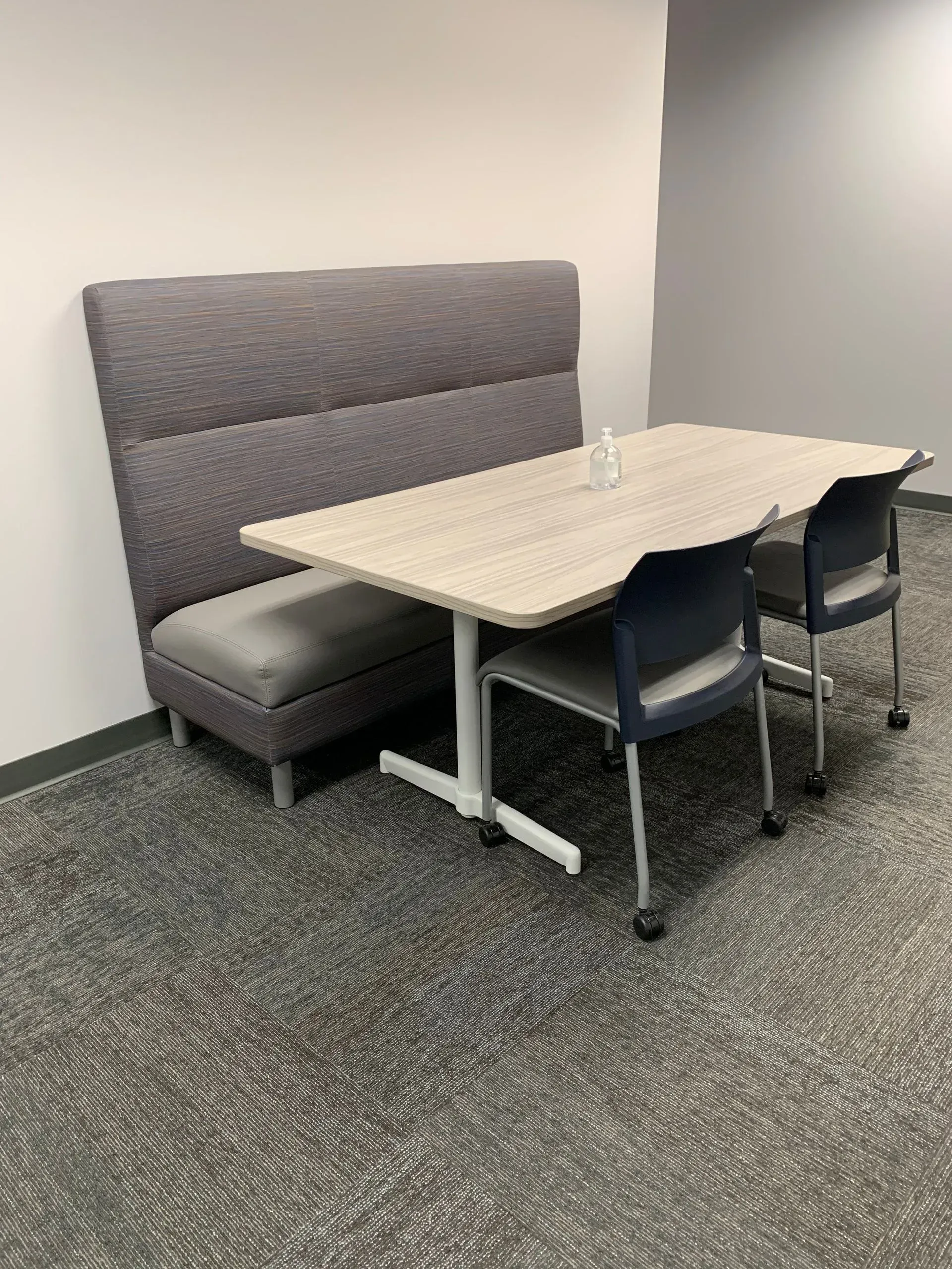 A booth with a gray cushioned back and table, two chairs, and a small glass on patterned carpet.