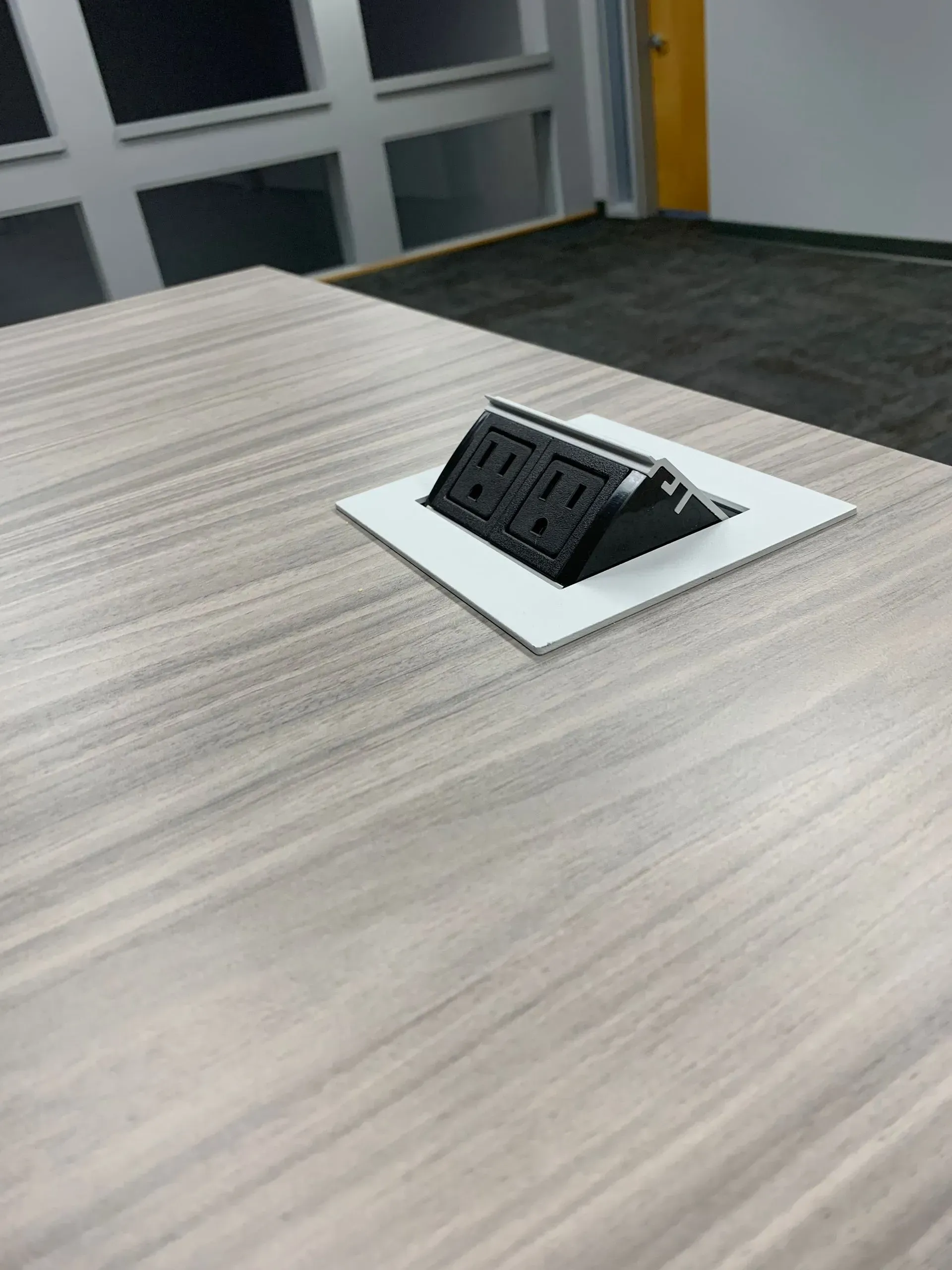 Gray table with built-in black power outlet, white trim. Office setting with windows and carpet in background.