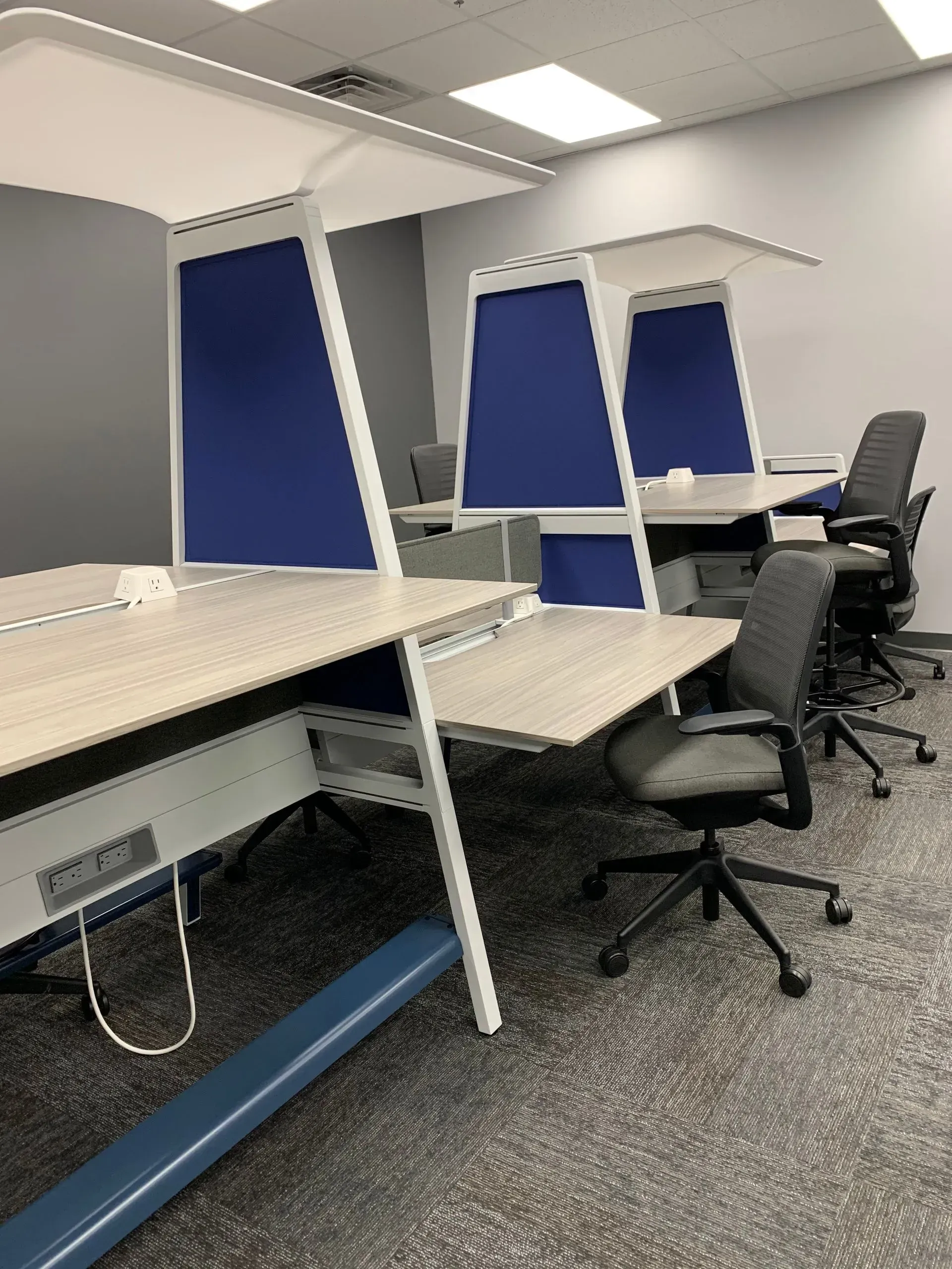 Office cubicles with gray and blue accents. Workstations with chairs and overhead lighting.