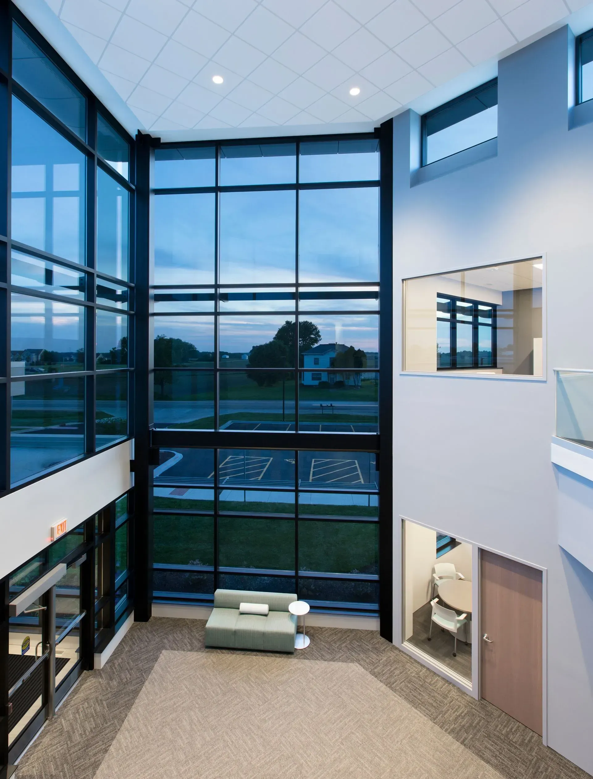 Modern lobby with large windows overlooking a green landscape. Gray walls, carpeted floor, and a small sofa.