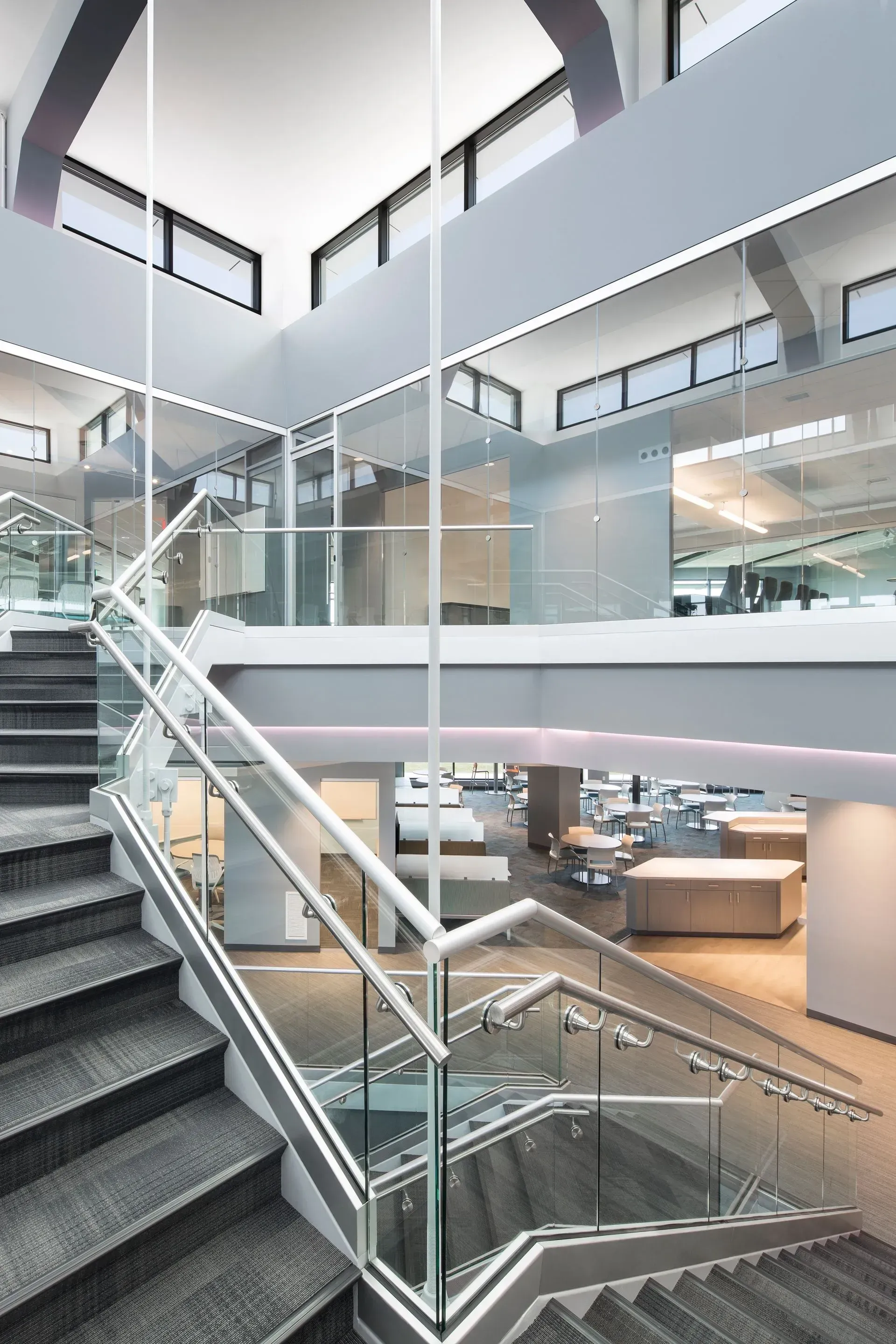 Staircase in modern building; gray carpeted stairs, glass railings, white walls, office space visible.