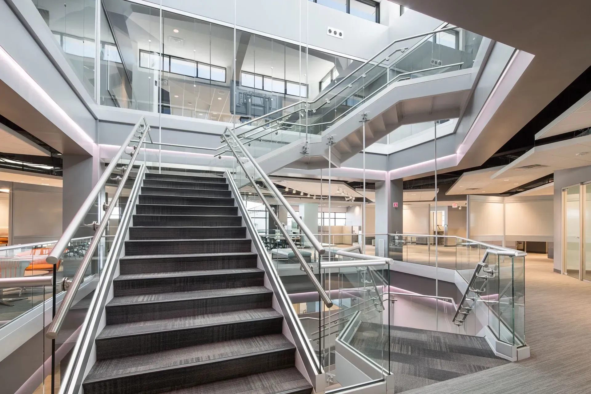 Staircase with glass railings in a modern building. Gray carpeted steps, white walls, and a multi-level design.