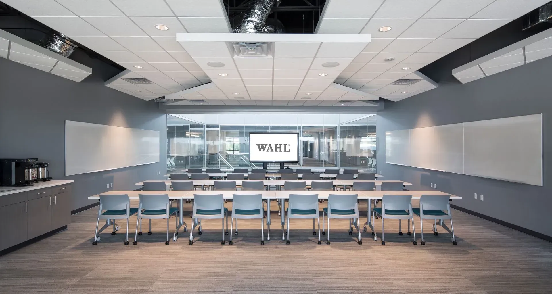 Conference room with long table and chairs, whiteboards, and a screen displaying a logo. Gray walls and carpet.