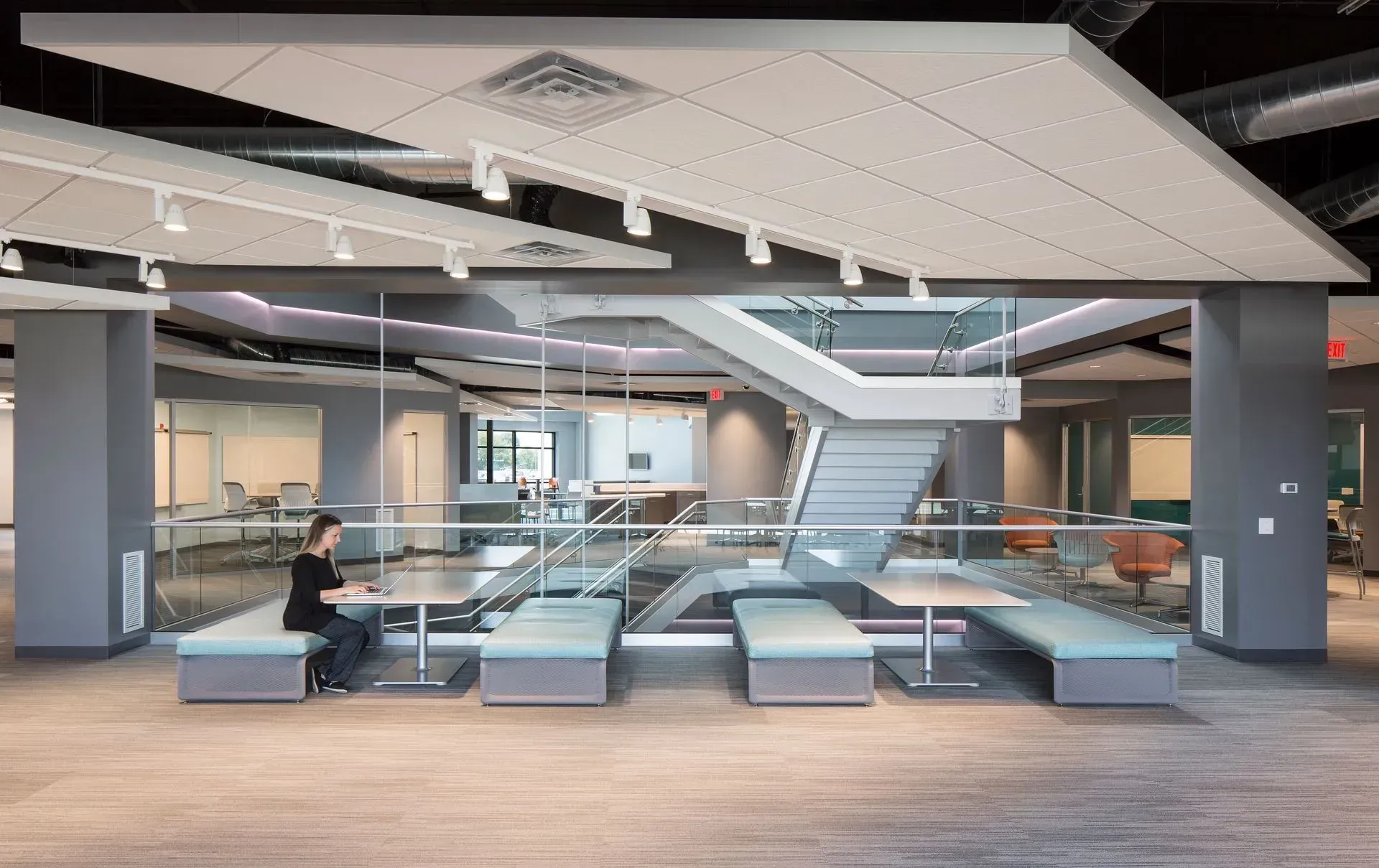 Woman seated at table in modern office space with a staircase, grey columns, and open workspaces.