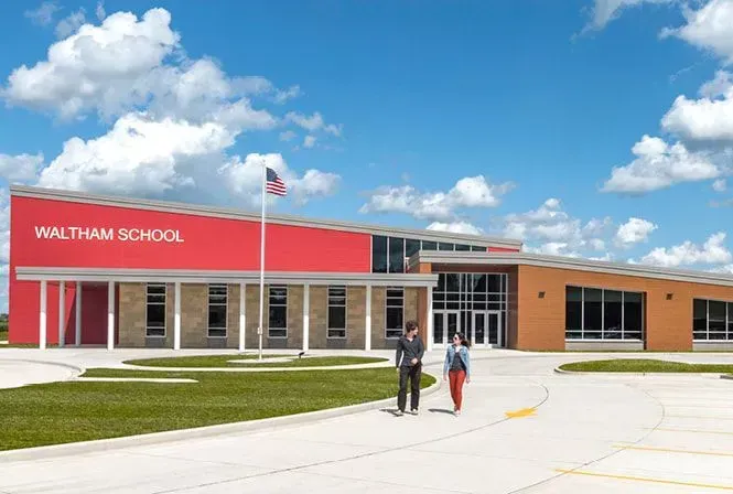Modern school building with red accent, two people walking on the sidewalk. American flag.