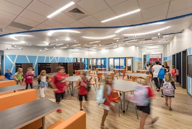 Children running in a brightly lit school cafeteria with tables and colorful walls.
