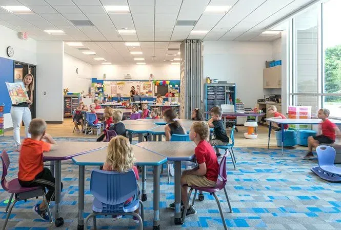 Classroom with children at tables, teacher showing a book. Blue patterned carpet, large window.