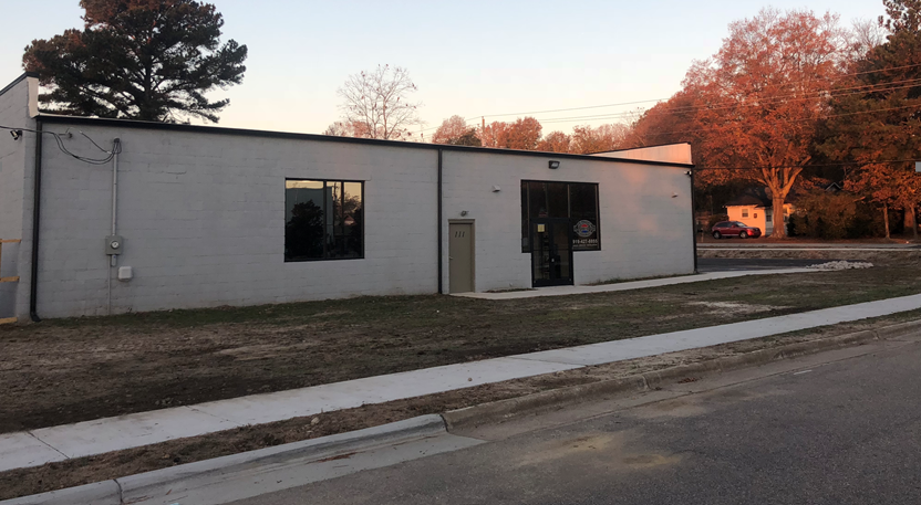 White building with a window and a door, set in a grassy area at dusk with trees in the background.