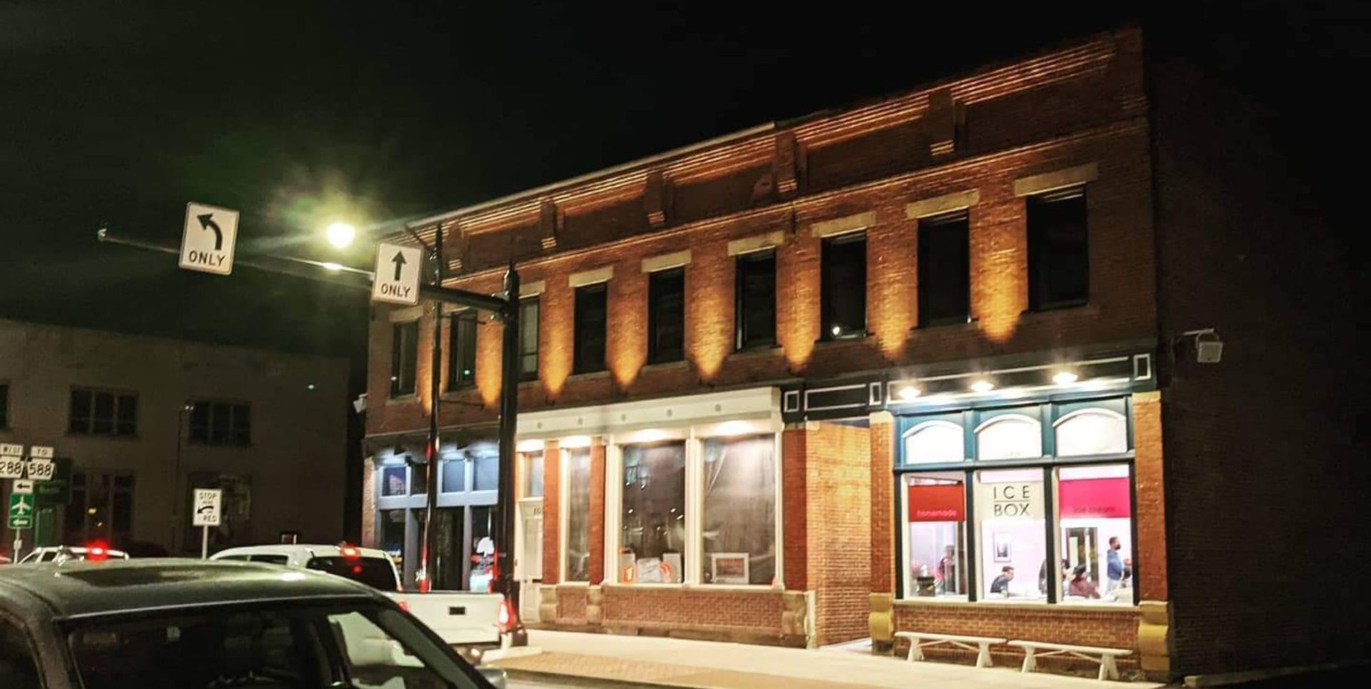 Nighttime street view of a lit brick storefront with large windows and parked cars outside
