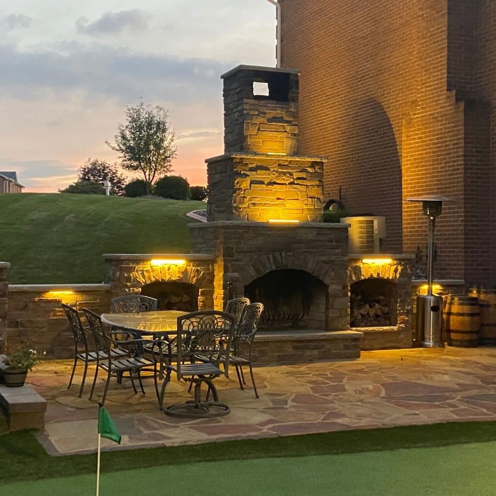 Outdoor stone patio with lit fireplace and metal tables beside a brick wall at dusk