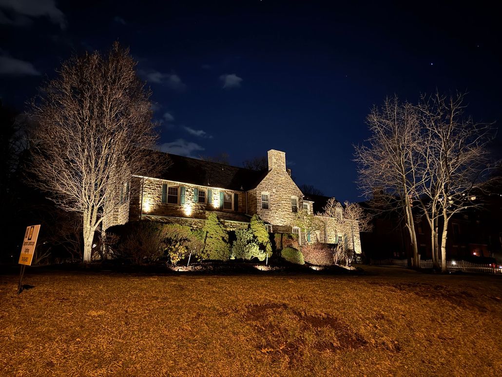 Illuminated stone house at night with bare trees and a lit lawn in front