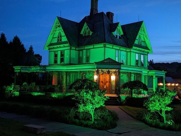 Victorian house lit green at night, with a glowing front porch and landscaped yard.