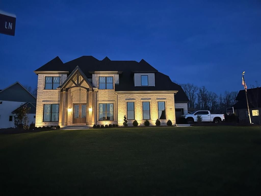 Two-story stone house lit at dusk with a dark roof and large lawn in front