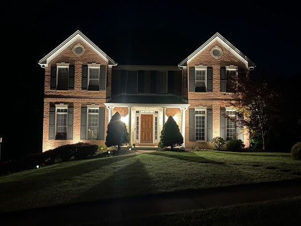 Two-story brick house at night, warmly lit front entrance, with two shrubs by the door and a dark lawn.