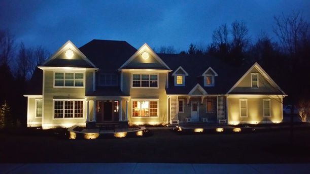 Large house at dusk with warm porch lights and lit dormers against a dark blue sky
