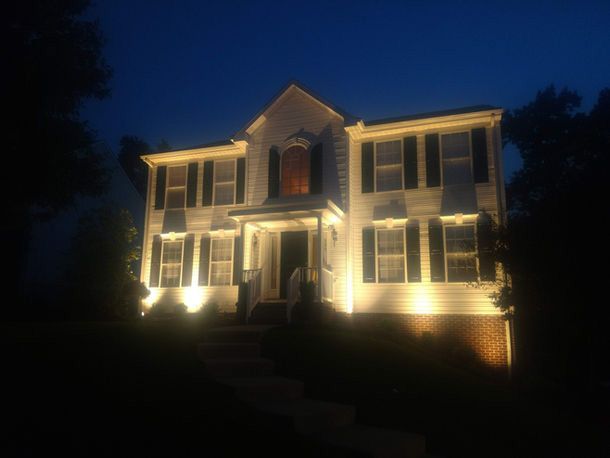 Two-story house at night, warmly lit with lights along the front and windows against a dark sky.