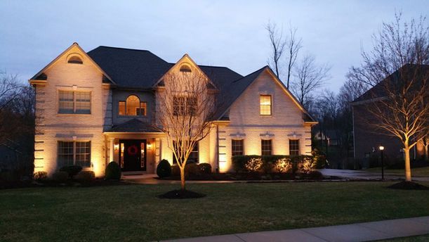 Large illuminated suburban house at dusk with lit windows, front lawn, and leafless trees