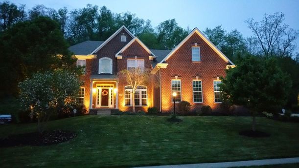 Illuminated brick house with warm lights at dusk, surrounded by trees and a dark lawn