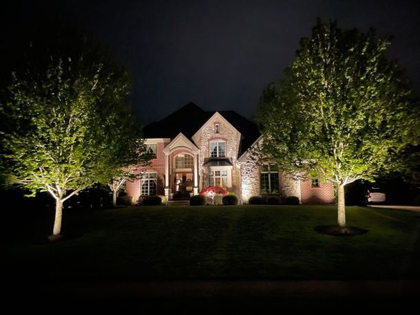 Well-lit brick house at night with two illuminated trees in the front yard