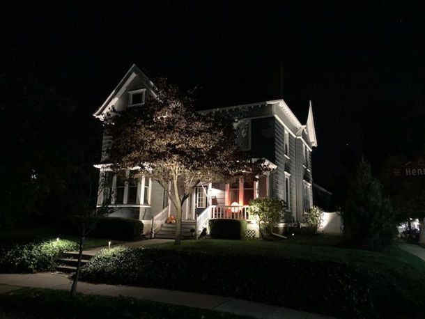 Lit Victorian-style house at night with bright porch lights and a small tree in the front yard