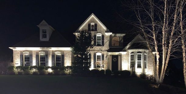 Illuminated suburban house at night with lit windows and bare trees in the yard