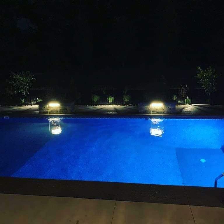 Nighttime view of a lit blue swimming pool with garden lights and trees in the background