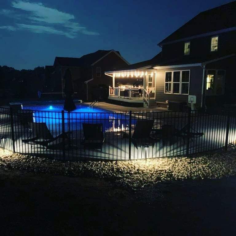 Backyard patio and glowing pool at night beside a lit house, enclosed by a black fence.
