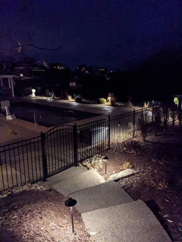 Nighttime view of a lit walkway and steps beside a black fence in a landscaped outdoor area