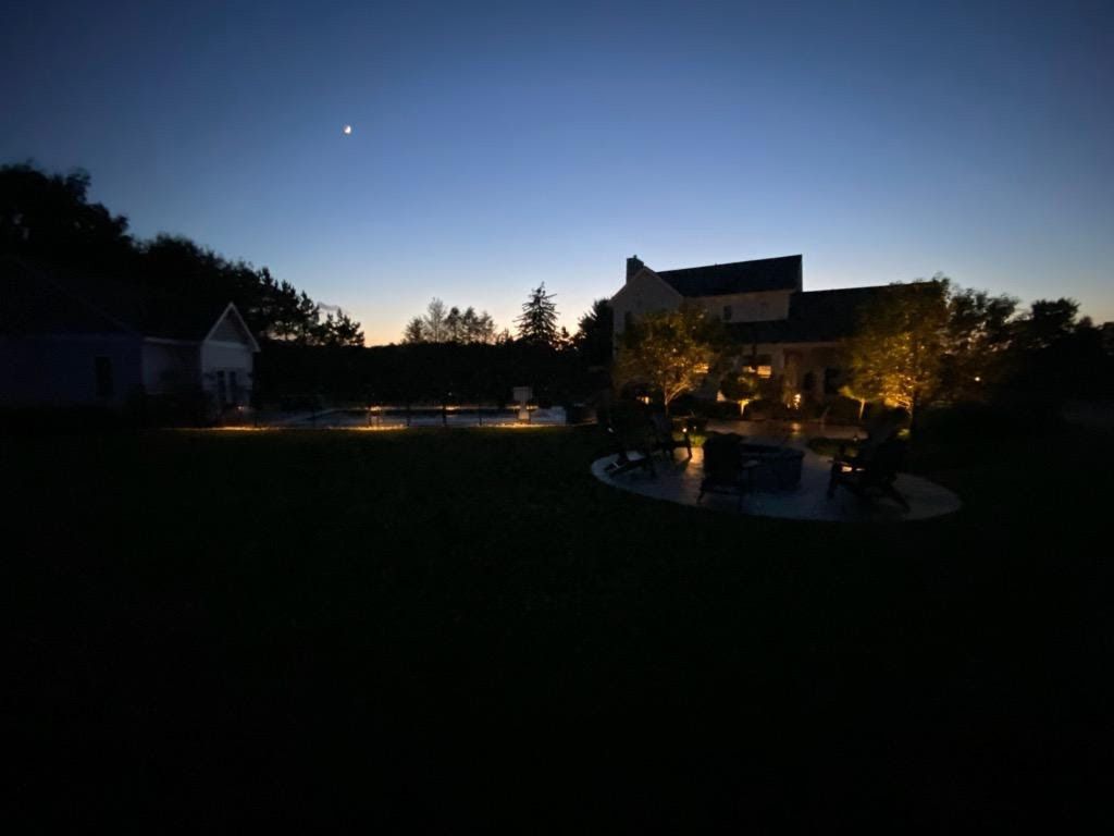 Dusk over a backyard with lit patio, dark trees, and a crescent moon in a blue sky