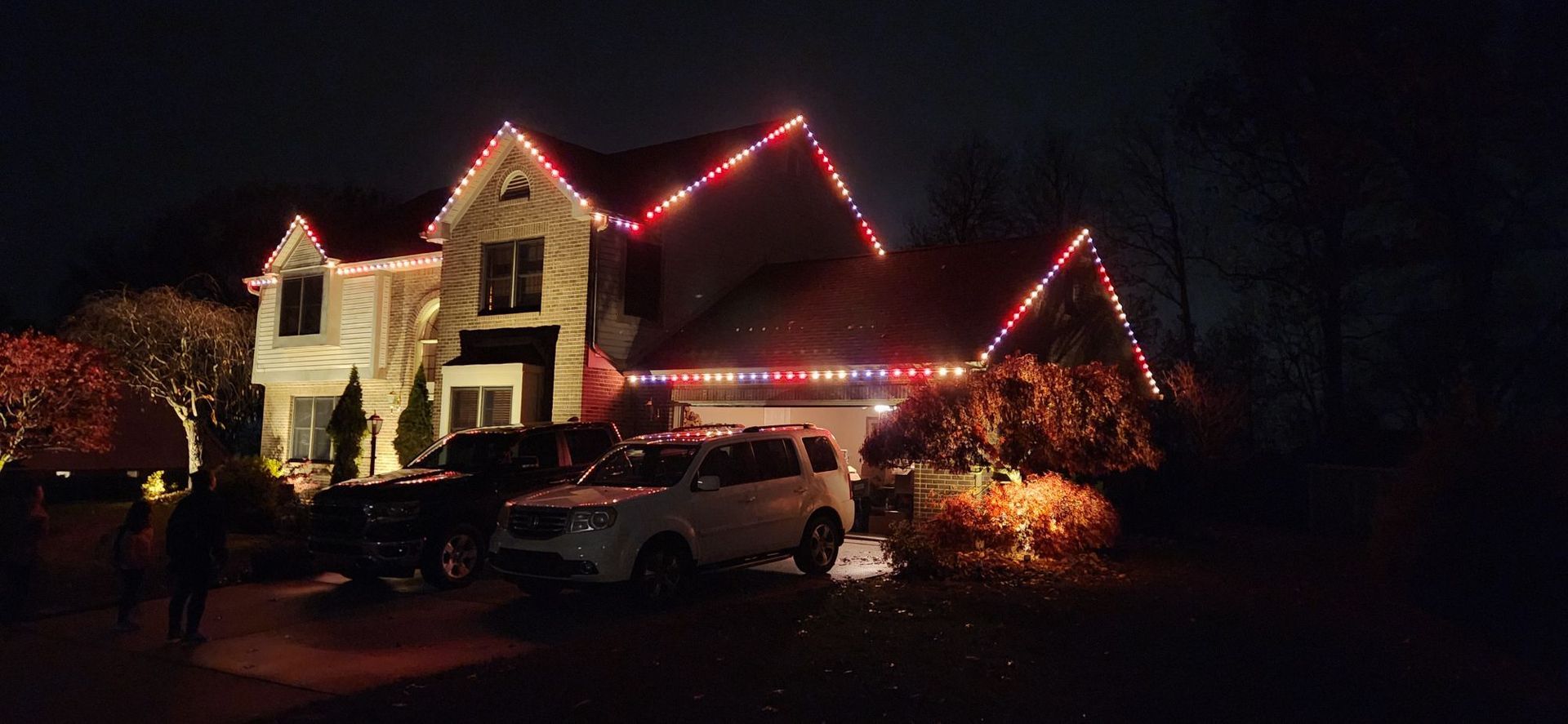 House lit with red holiday lights at night, with parked cars in the driveway