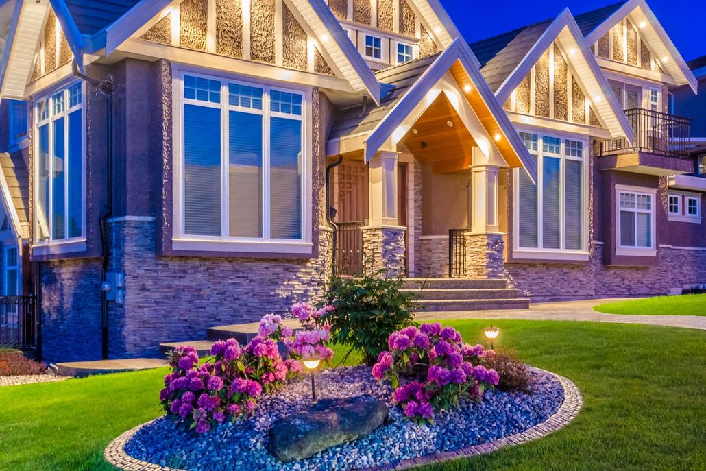 Luxury home exterior at dusk with warm lights and a flower bed in front