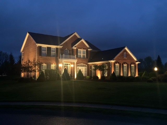 Large brick suburban house lit warmly at dusk, with a dark blue evening sky and manicured lawn.