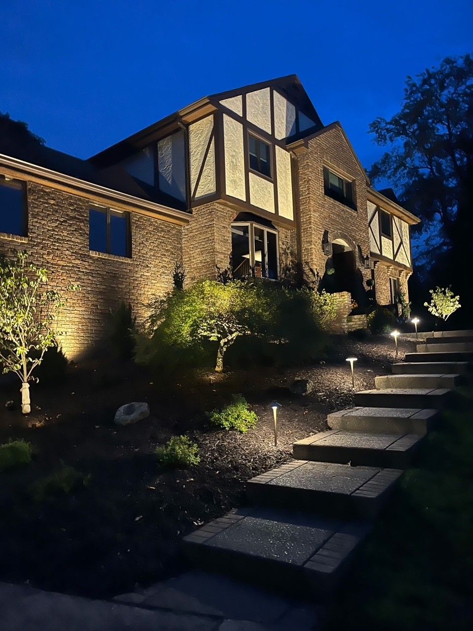 Stone house at dusk with lit windows and a stepped garden path under a deep blue sky