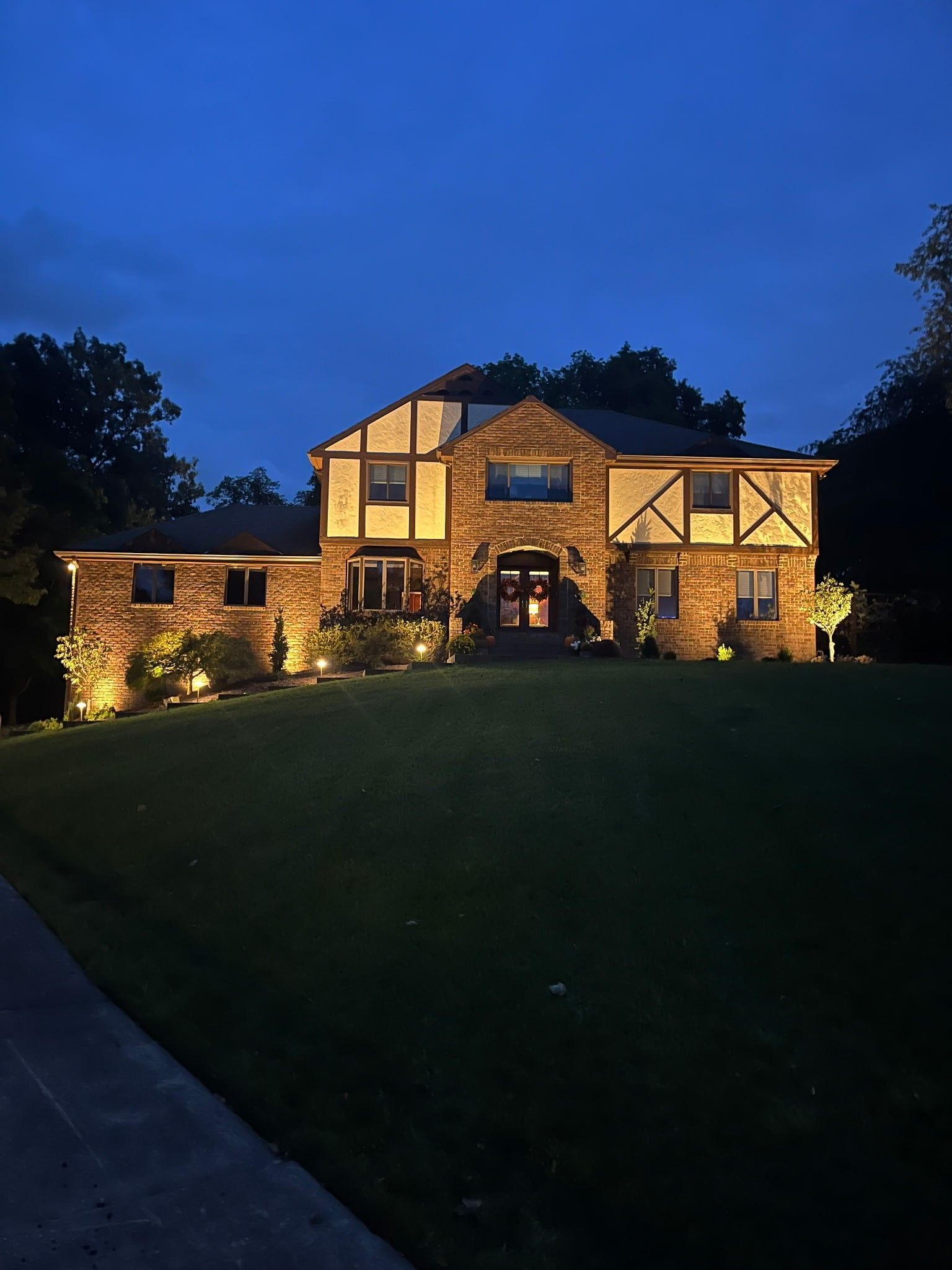 Two-story stone house lit warmly at dusk, with a dark lawn and deep blue evening sky