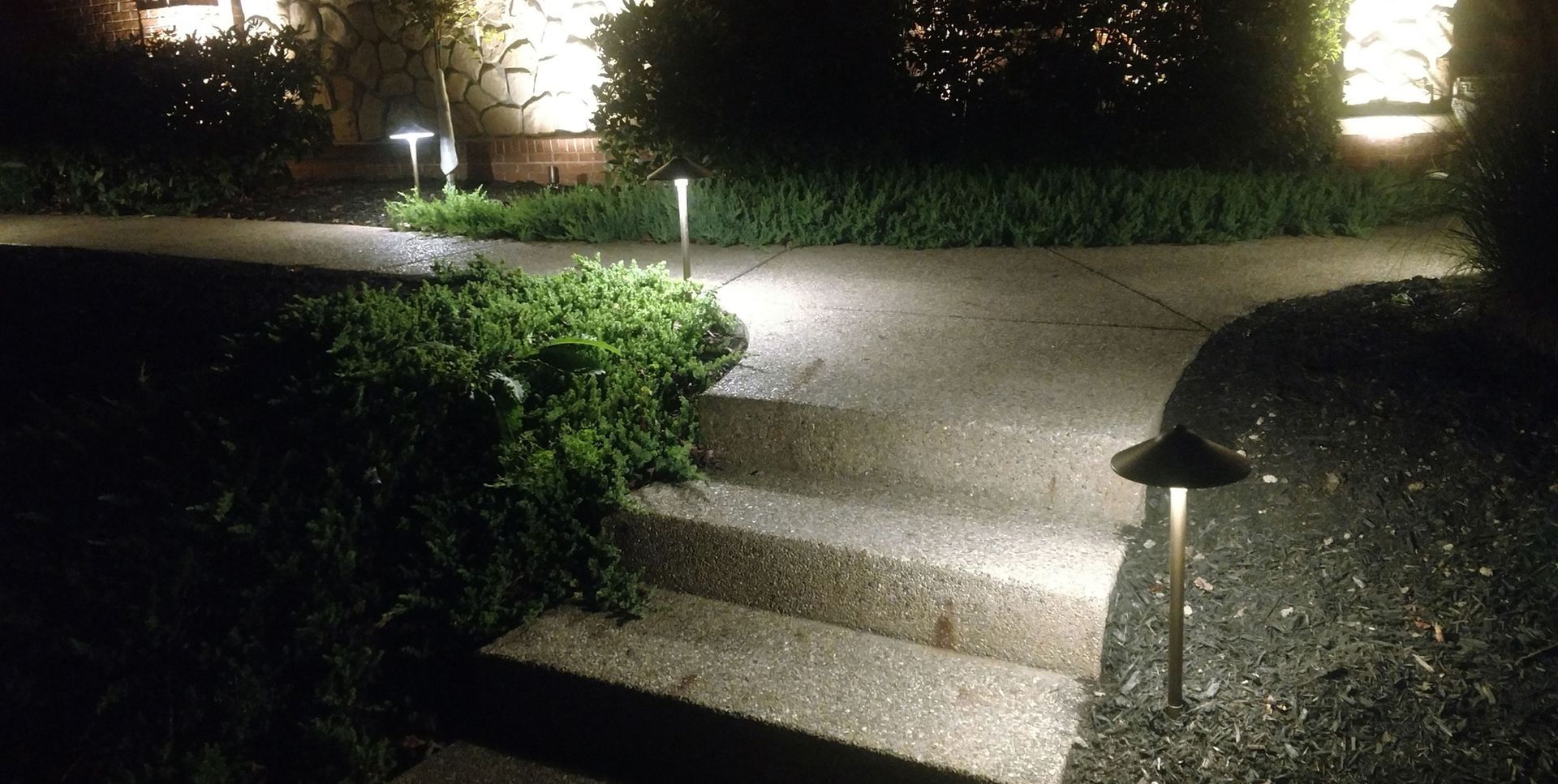 Dimly lit garden path with stone steps, hedges, and small lamp posts at night