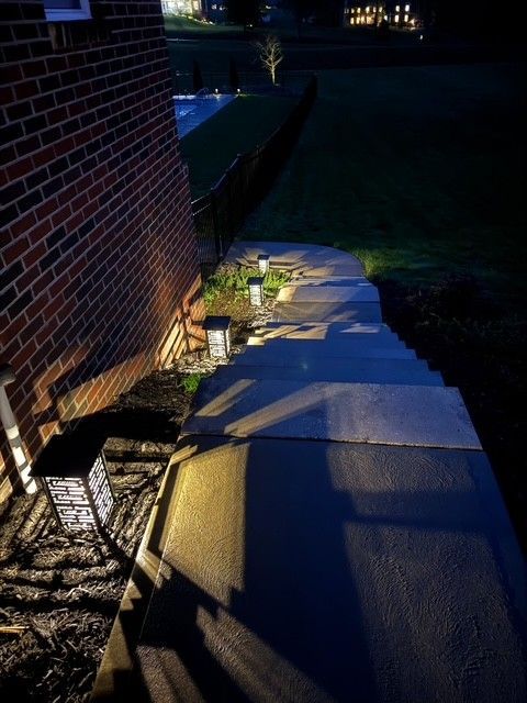 Lit brick walkway beside a house at night, with small landscape lights and shadows on the path. 